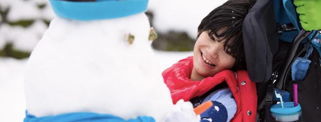 A child with cerebral palsy sits in a wheelchair and smiles while looking at a snowman