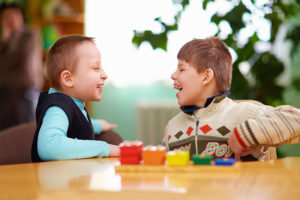 Two young children with cerebral palsy laughing and playing with colorful blocks at a table.