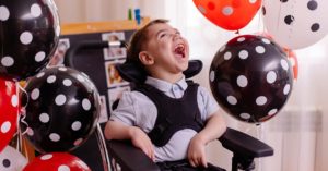 A boy with cerebral palsy smiles as he sits in a wheelchair surrounded by balloons.